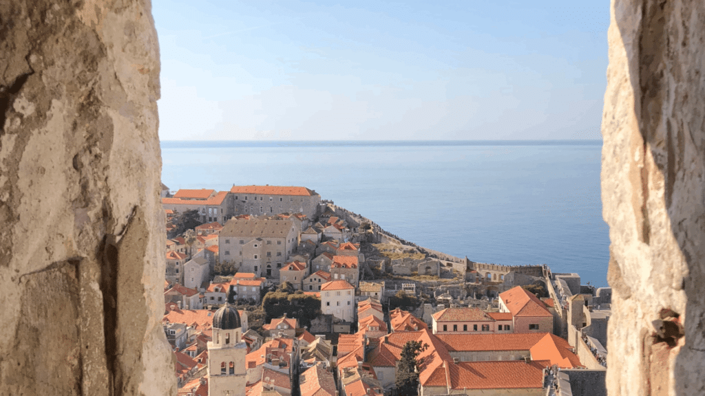 View of Dubrovnik Old Town through its historic stone walls, with red-tiled rooftops and the Adriatic Sea beyond.